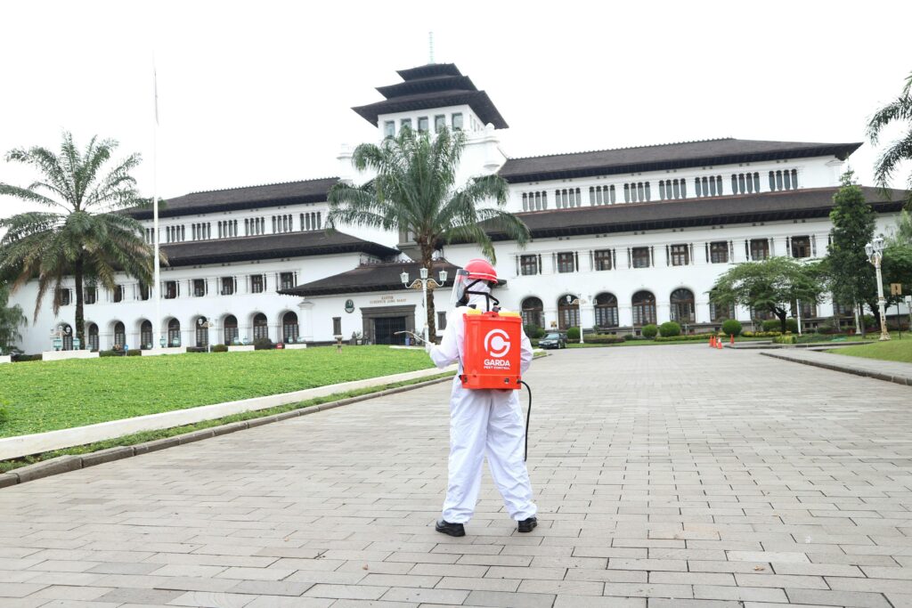 A person in protective clothing disinfects an area in front of a historic building with palm trees.